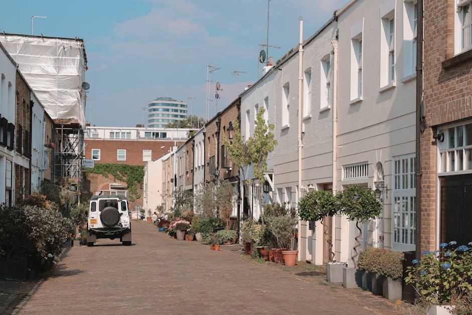 A narrow cobblestone street in a residential area lined with closely spaced terraced houses. Each house has a small front garden or potted plants, with some featuring well-maintained shrubs, small trees, and flowering plants in terracotta pots. The facades of the houses are a mix of white and red brick, with windows and doors varying in style. On the left side, a white van is parked, and some houses show signs of recent garden maintenance, such as trimmed hedges and tidy flower beds. Overhead, the sky is clear with a few wispy clouds, and in the background, a modern multi-storey building is visible, contrasting with the traditional row of houses. This scene reflects a peaceful urban neighborhood with signs of ongoing outdoor care and garden maintenance, aligning with landscaping work and lawn care themes related to the services provided by Gardeners Streatham, as referenced in the page about same day gardening availability and costs.