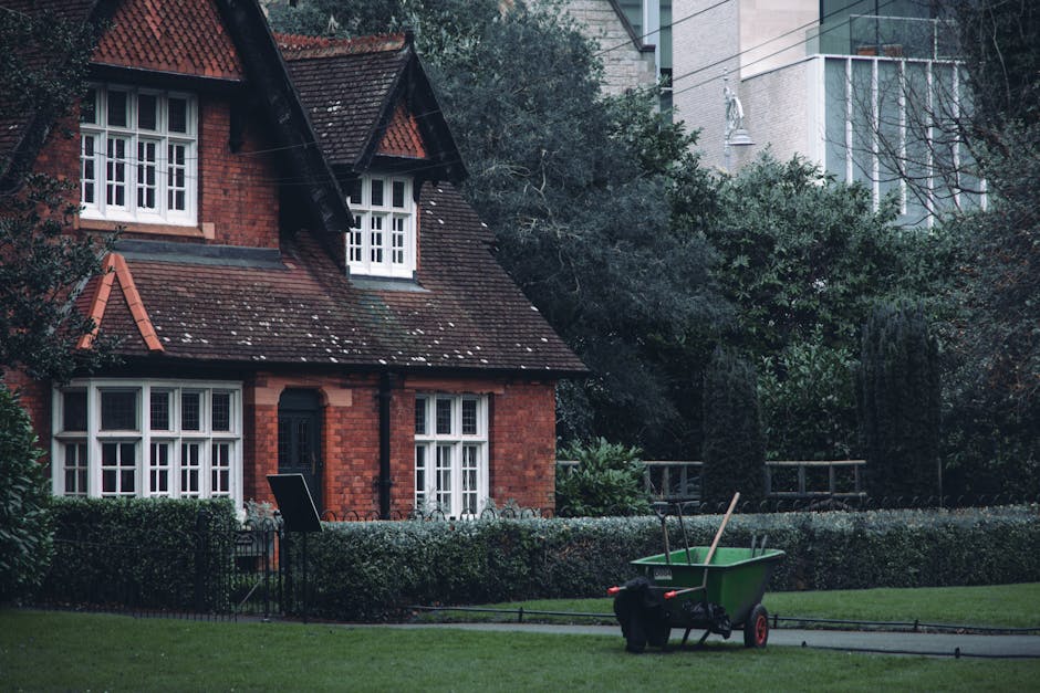 A red brick house with a sloped tiled roof and white-framed windows surrounded by a well-maintained garden. The garden features a neatly trimmed hedge bordering a lush green lawn, with some trees and shrubs providing natural privacy. In the foreground, a green wheelbarrow with red handles and a black bag inside is parked on a paved pathway, alongside rakes and garden tools, indicating ongoing garden maintenance or landscaping work. The scene is lit by natural daylight, creating a calm atmosphere suitable for outdoor lawn care and hedge trimming activities, with the emphasis on the organized outdoor space associated with professional gardening services from Gardeners Streatham.