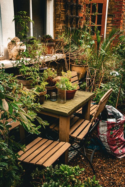 A cozy urban garden corner featuring a weathered wooden table surrounded by matching wooden benches and chairs. The table is decorated with potted plants, including succulents and leafy foliage, along with a glass bottle and small decorative items. Lush greenery, including potted palms, shrubs, and trailing plants, fills the space, creating a densely vegetated environment. The garden is situated next to a brick house with a large window and a wooden door, enhancing the intimate atmosphere. The ground is covered with small gravel, and there are various garden tools and a large fabric storage bag visible, indicating ongoing garden maintenance and care. The scene is bathed in natural daylight, emphasizing the vibrant green hues and textured surfaces of the plants and garden furniture. This setting reflects a well-tended small garden, maintained by Gardeners Streatham, highlighting the themes of outdoor landscaping and garden upkeep.